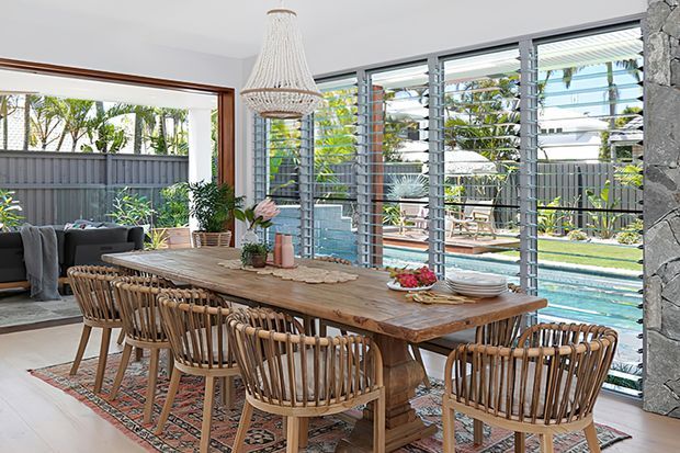 Bright dining area with a large rustic timber dining table, woven wooden dining chairs, patterned rug, and a woven pendant light. Full-height louvred glazing opens views to a swimming pool and landscaped outdoor area, while a dark stone feature wall and warm timber window framing add texture.