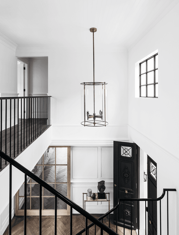 A bright, double-height residential entry hall viewed from a staircase landing. The space features crisp white walls and ceiling, black metal balustrades, dark herringbone timber flooring, a tall suspended lantern pendant, paneled wall detailing, a black decorative front door, and large gridded windows. A console table and sculptural decor sit in the foyer below.