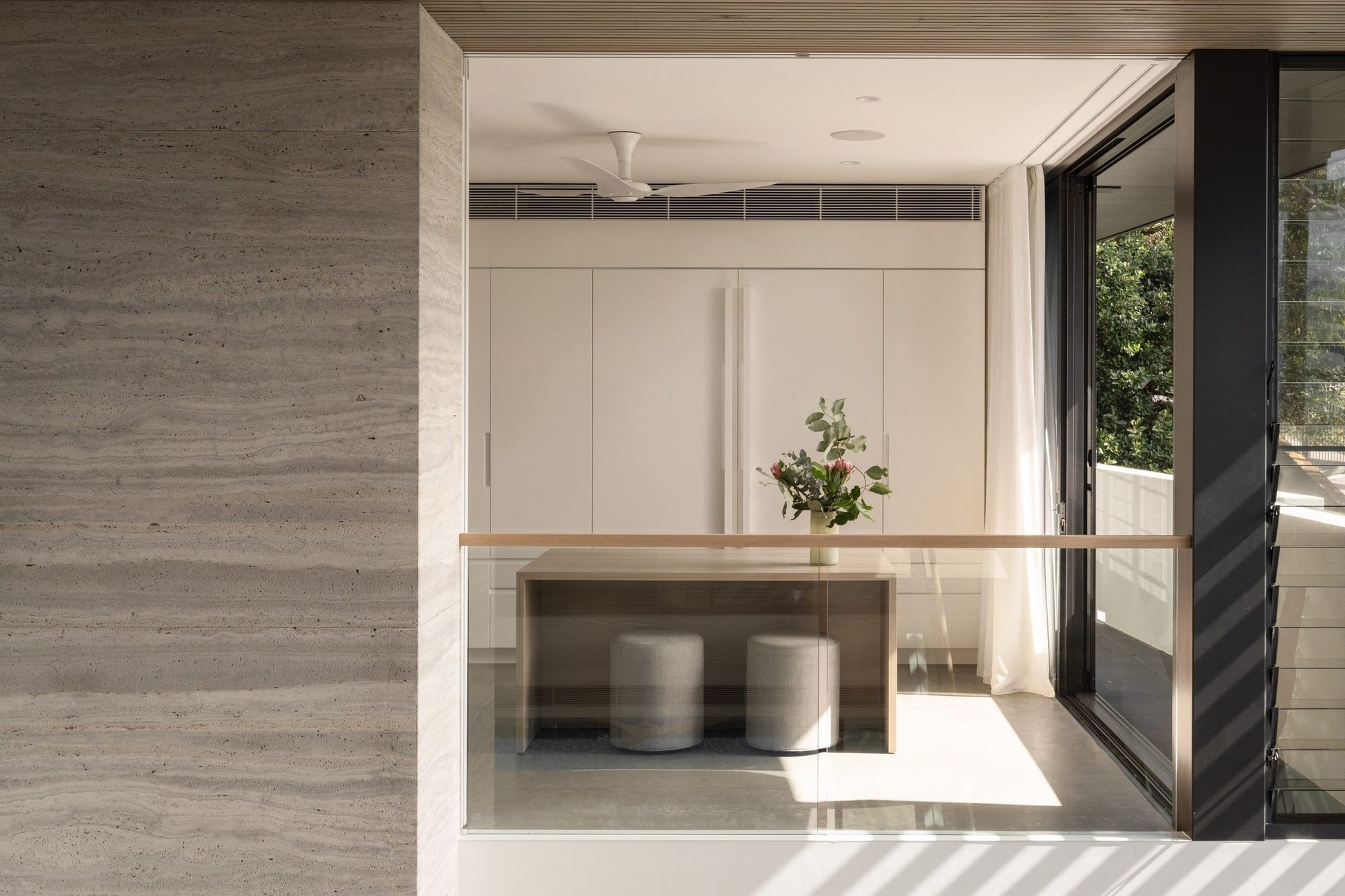 Decade House - Kitchen. Breakfast bar beside a travertine wall, with white cabinetry and garden views.