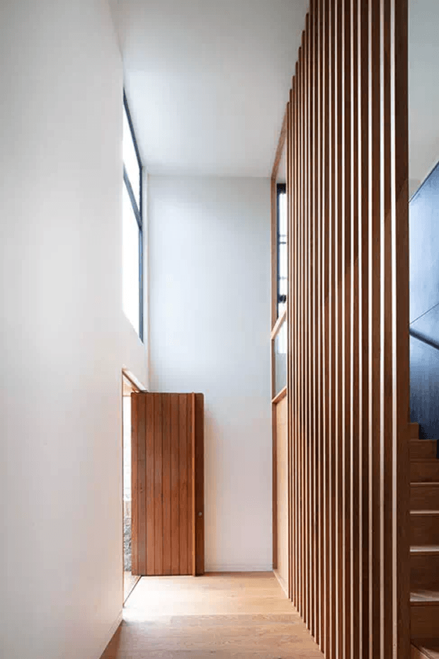 Minimalist residential hallway with warm timber flooring, a vertical timber slat screen forming a stair balustrade or partition, a timber door, white plaster walls, and strong natural light from narrow windows. The stairway is partially visible behind the slatted screen.