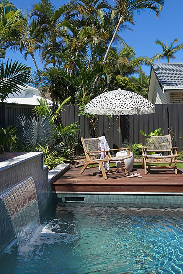 A tropical outdoor pool area with a raised timber deck, tiled pool edge, built-in water feature, and two woven lounge chairs beneath a patterned parasol. Dense palms and lush planting surround the dark privacy fence, with neighbouring rooflines visible beyond.
