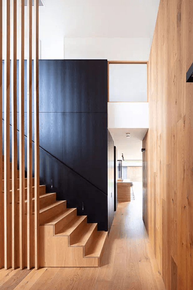 A bright, contemporary residential hallway centered on a timber staircase. Vertical timber battens form a screen along the stair edge, while a dramatic matte-black stair wall and dark balustrade contrast with warm timber flooring and full-height timber wall panelling. The corridor extends toward a brighter living or kitchen area, with a partial upper-level opening visible.