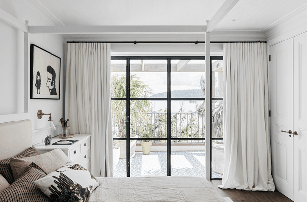 Bright, refined bedroom with white panelled walls and ceiling, a partially visible upholstered bed, neutral layered bedding, and full-height cream curtains framing black steel-style glazed doors. The glazing opens to a tiled terrace or balcony with railing and greenery beyond. A monochrome artwork, wall-mounted reading light, and white paneled double doors complete the calm coastal-classic interior.
