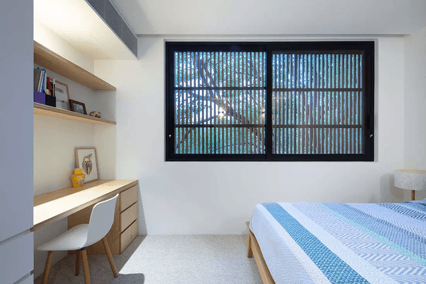 Bright, compact bedroom with a built-in timber study desk and open shelving along one wall. A white molded chair sits at the desk, while a bed with blue-and-white patterned bedding occupies the right side. The room has white walls, light carpeting, a large black-framed window with vertical privacy screening, and a clean contemporary feel.