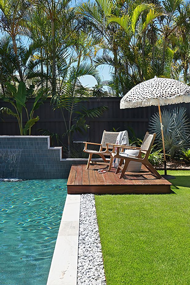A sunny landscaped backyard pool area with a tiled swimming pool, raised timber deck, mid-century-inspired outdoor lounge chairs, a fringed patterned parasol, tropical palms, dark fencing, and manicured lawn. White decorative gravel borders the pool and deck, creating a resort-like outdoor setting.