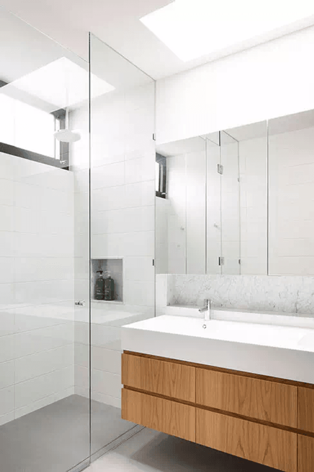 Bright contemporary bathroom with a frameless glass walk-in shower, white tiled walls, large mirrored cabinetry, a white integrated vanity basin, and warm timber floating drawers. A skylight and high-level glazing provide abundant natural light.