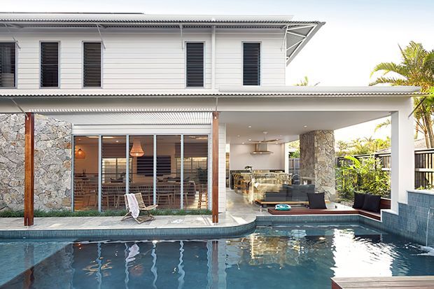 Bright exterior view of a two-storey white-clad residence with a covered outdoor entertaining area, swimming pool, and open-plan living and kitchen spaces visible through glazing. The facade combines horizontal weatherboard cladding, stone feature walls, timber posts, dark-framed windows, and a corrugated metal roof.