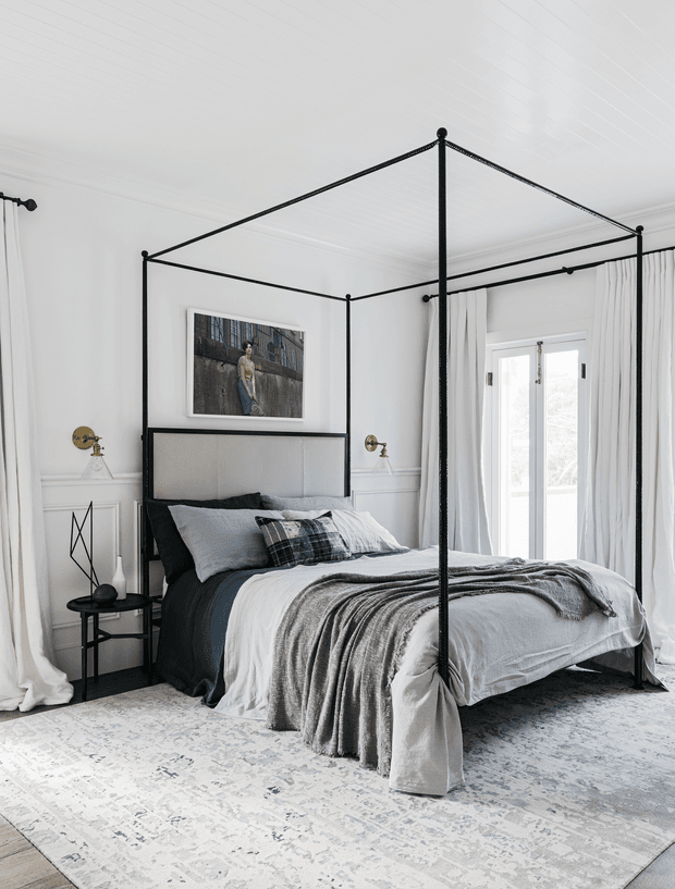 A bright, refined bedroom featuring a black metal four-poster canopy bed with a light upholstered headboard, layered white and charcoal bedding, and a draped grey throw. White walls with decorative wainscoting, tall white curtains, brass wall sconces, framed artwork, a black side table, and a pale patterned rug create a contemporary classic monochrome scheme. French doors provide abundant natural light.