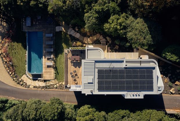 Aerial view of a contemporary flat-roofed building set among mature trees, with a large rooftop solar-panel array, rooftop terrace or deck areas, and an adjacent narrow swimming pool with timber decking and landscaped stone edging. A paved driveway runs alongside the property.