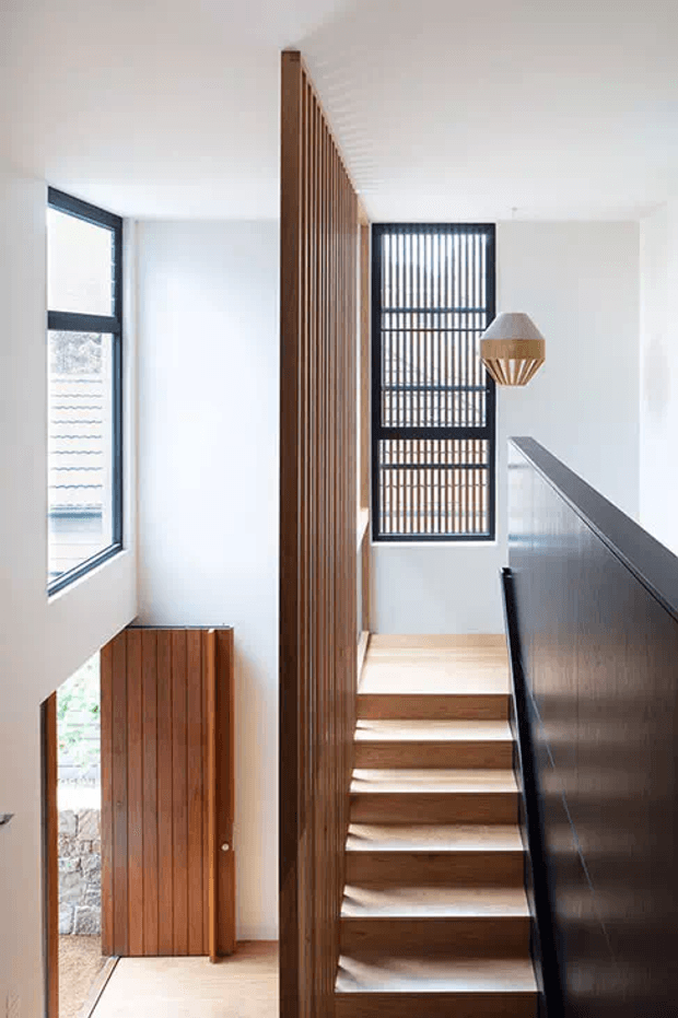 Bright modern stairway with warm timber treads and a tall vertical timber slat divider and balustrade. White walls and ceiling contrast with dark-framed louvred windows, a black stair-side wall, and a sculptural pendant light. A timber entry door is visible below the stairs.
