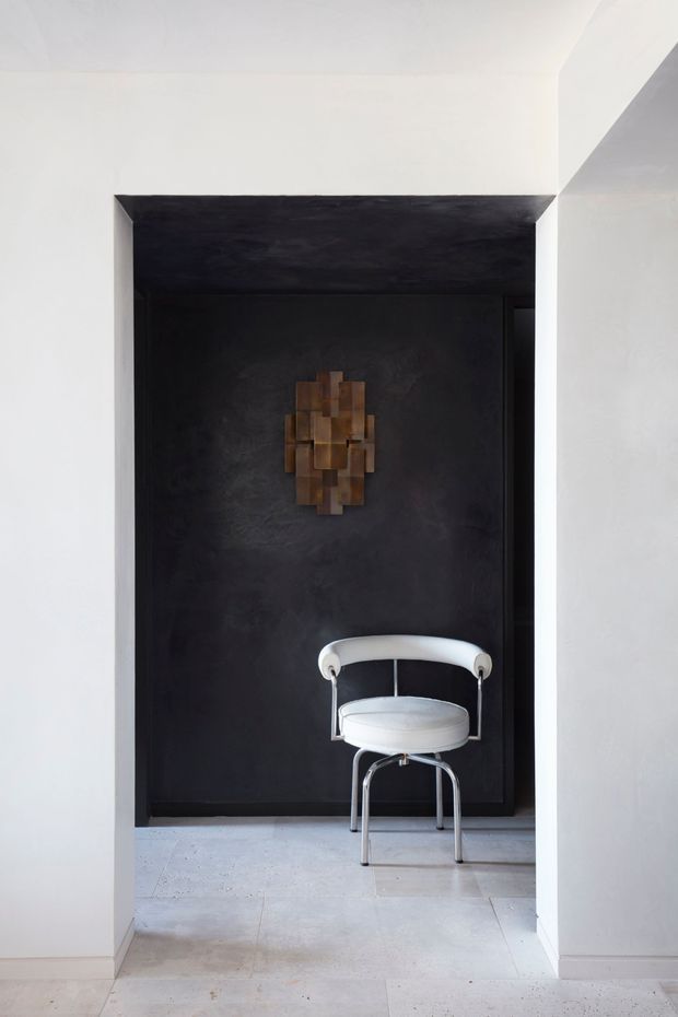 A minimalist interior view through a white-walled opening into a dark, recessed architectural alcove. The alcove has a black textured feature wall and ceiling, a sculptural brown wall artwork, and a distinctive white tubular lounge chair with chrome-finished legs. Pale stone or concrete-look floor tiles continue through the space, creating strong contrast between the bright circulation area and dark niche.