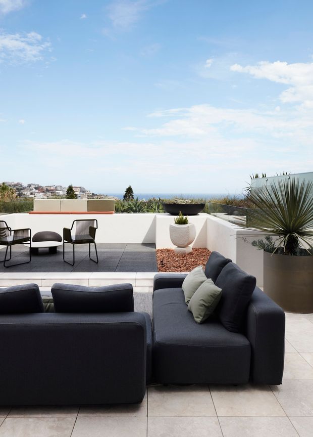 A bright rooftop or elevated outdoor terrace overlooking the sea and a coastal residential neighbourhood. The foreground contains a dark charcoal outdoor sectional sofa with neutral cushions, while a separate dining area has black metal-framed chairs and a low table. White rendered parapet walls, pale large-format tiled paving, gravel planter beds, and sculptural drought-tolerant plants create a clean contemporary coastal setting under a blue sky.