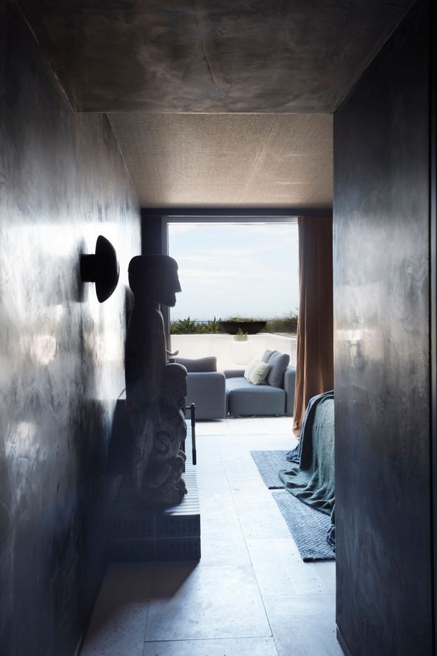 A dark, narrow interior passage or bedroom entry opens toward a bright outdoor terrace and ocean horizon. The space has deep charcoal textured walls and ceiling, large pale rectangular floor tiles, a low built-in or sculptural bench along the left wall, and a dark upholstered bed or seating edge on the right. At the far end, a grey lounge chair and ottoman sit beside a large opening, with sheer coastal light and vegetation beyond. The atmosphere is dramatic, intimate, and strongly contrasted with the bright exterior.