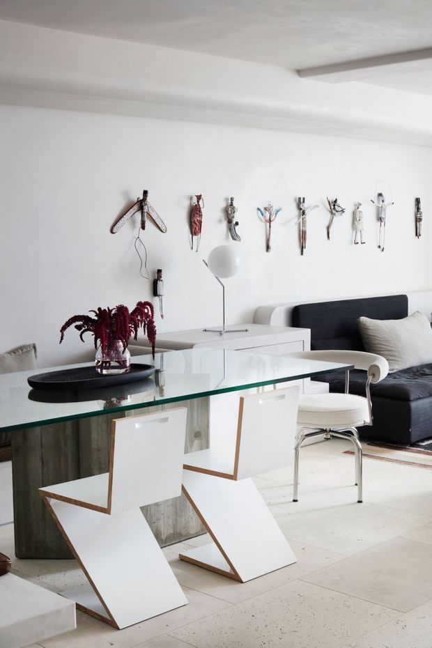 Bright contemporary open-plan interior showing a dining area in the foreground and a living room behind it. A clear glass dining table with sculptural white geometric chairs sits over a pale tiled floor. The room includes a dark upholstered sofa, white armchair, minimalist floor lamp, white cabinetry or console, and a wall-mounted collection of sculptural objects. The palette is predominantly white and light grey with black, dark charcoal, and burgundy accents. The architecture is simple and refined, with a white ceiling and exposed beam.