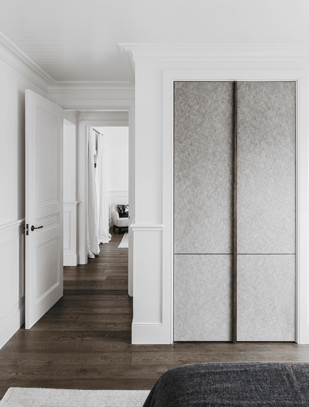 A refined, light-filled residential hallway with white painted wall panelling, decorative cornices and skirting, dark timber flooring, and a large built-in wardrobe or storage unit with textured grey fabric-fronted doors. An open panelled door leads toward a softly furnished bedroom or sitting area in the background. The styling is restrained, elegant and monochromatic with warm timber contrast.