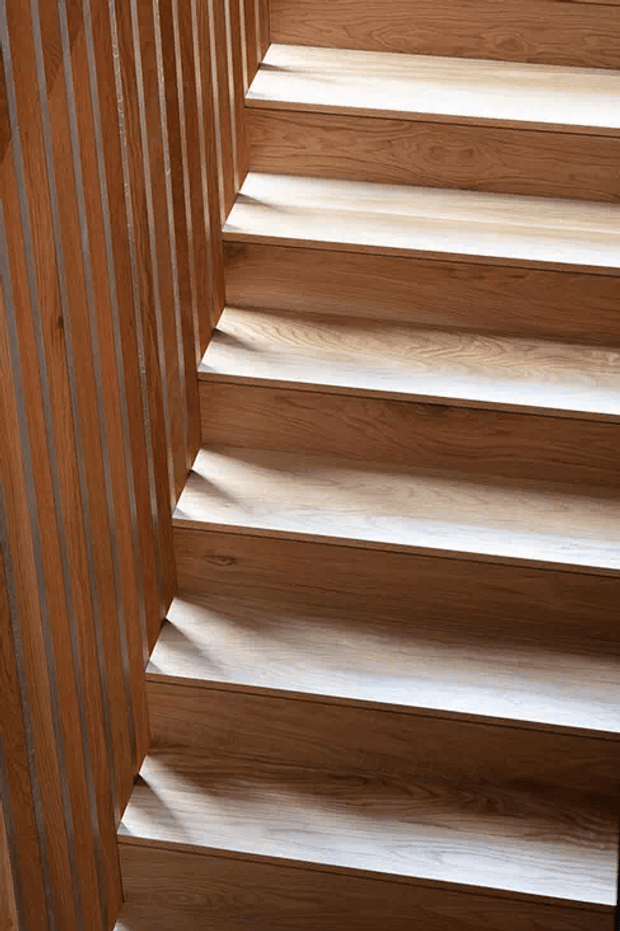 Close-up view of a timber staircase with broad wooden treads and risers, bordered by a vertical timber slat wall or balustrade. The natural wood grain and warm brown finish dominate the image.