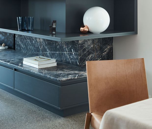 A refined contemporary interior featuring a dark built-in storage and display unit with a dramatic veined stone benchtop and matching splashback. A white spherical lamp, glass vessels, books, and a warm tan leather chair sit against the deep charcoal joinery, with a light stone or terrazzo floor and a partially visible dining table in the foreground.