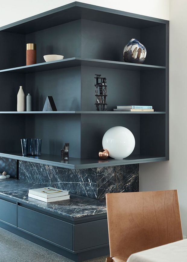 A sophisticated dark built-in cabinetry and shelving unit occupies the corner of a dining or living area. The matte charcoal joinery features open display shelves styled with ceramics, glassware, books, sculptures, and decorative objects. Below, a dramatic dark marble or natural stone benchtop and splashback wrap around low cabinetry, with a tan leather dining chair in the foreground and part of a light dining table visible. The composition has a refined, moody contemporary feel.