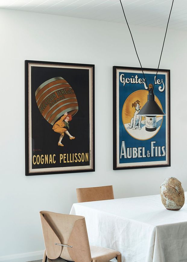 Bright, minimalist dining area with a white-clothed rectangular table, sculptural wooden dining chairs, and two framed vintage cognac advertising posters on a white wall. A black pendant light hangs over the table, while a textured stone-like decorative object provides a natural accent.