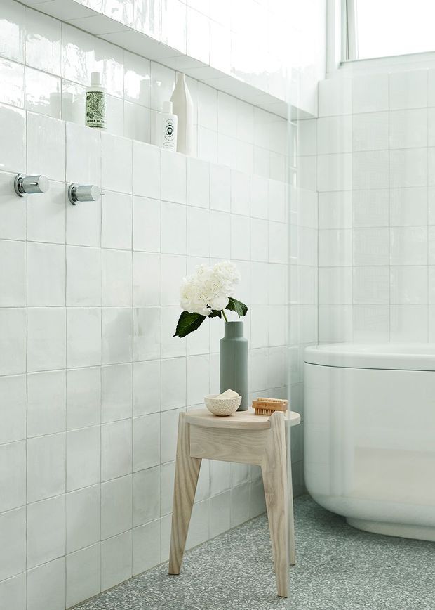 Bright, serene bathroom with white square wall tiles, a freestanding white bathtub, chrome wall fixtures, and a speckled terrazzo-style floor. A pale timber stool holds a vase, bowl, and grooming accessories, while recessed tiled shelving and a high window add practical storage and natural light.