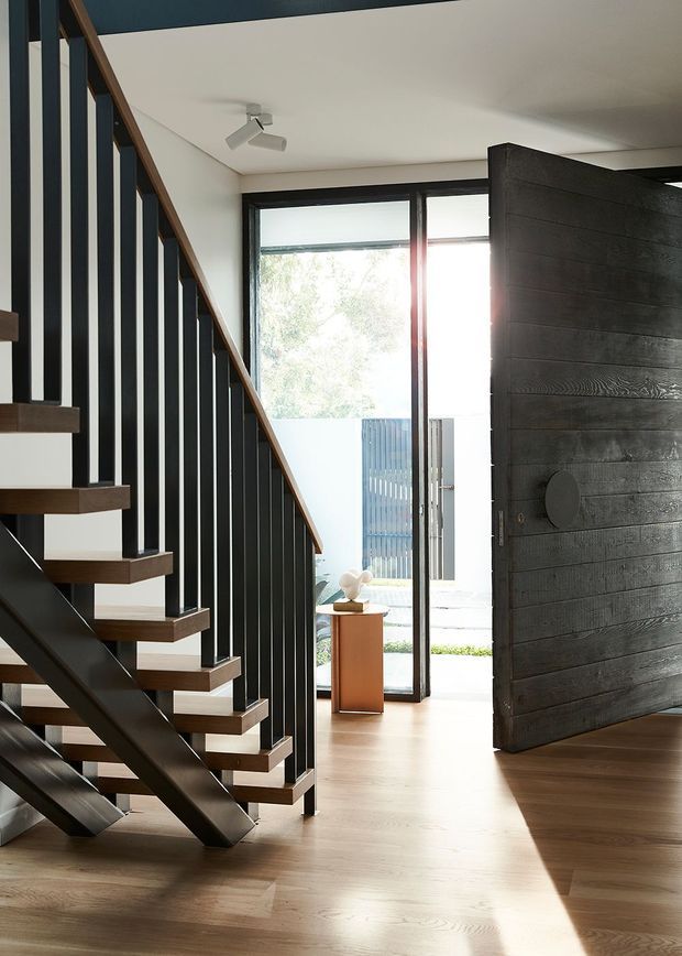 A bright contemporary entryway featuring an open-riser staircase with dark metal balustrades and timber treads, pale timber flooring, and a large dark timber pivot front door. Full-height glazing opens toward an outdoor entry area, bringing in strong natural light.