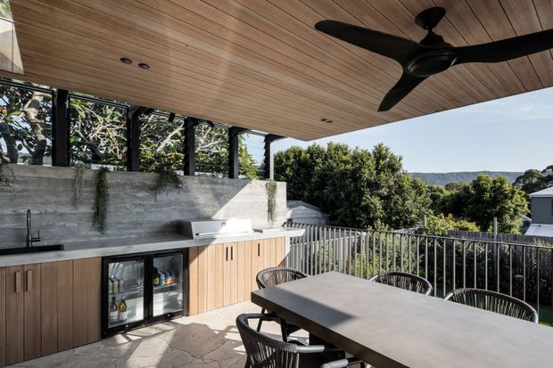 A covered outdoor entertaining area with a built-in kitchenette, concrete backsplash and benchtop, timber cabinetry, under-counter beverage fridge, dining table and chairs. The space has a timber-lined ceiling, large black ceiling fan, metal balustrade and views toward a leafy suburban landscape.