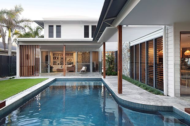 Contemporary residential courtyard viewed from beside a narrow swimming pool, with white weatherboard-clad volumes, timber posts and vertical screening, extensive louvred glazing, a covered outdoor entertaining area, and a raised lawn edge. The pool and connected indoor-outdoor spaces are the dominant features.
