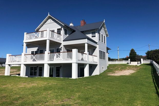 Exterior view of a large multi-level coastal-style house with white horizontal weatherboard cladding, dark pitched gabled roofs, multiple elevated verandas and balconies with white railings, and open lower-level spaces. The house sits on a broad grassy site under a clear blue sky.
