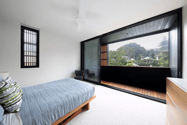Bright contemporary bedroom with a low timber bed, pale blue quilt, white walls, carpeted floor, and a large black-framed glazed opening leading to a timber-decked enclosed balcony or terrace. A black-framed barred window, ceiling fan, and light timber drawers complete the room. Greenery is visible beyond the glazing.