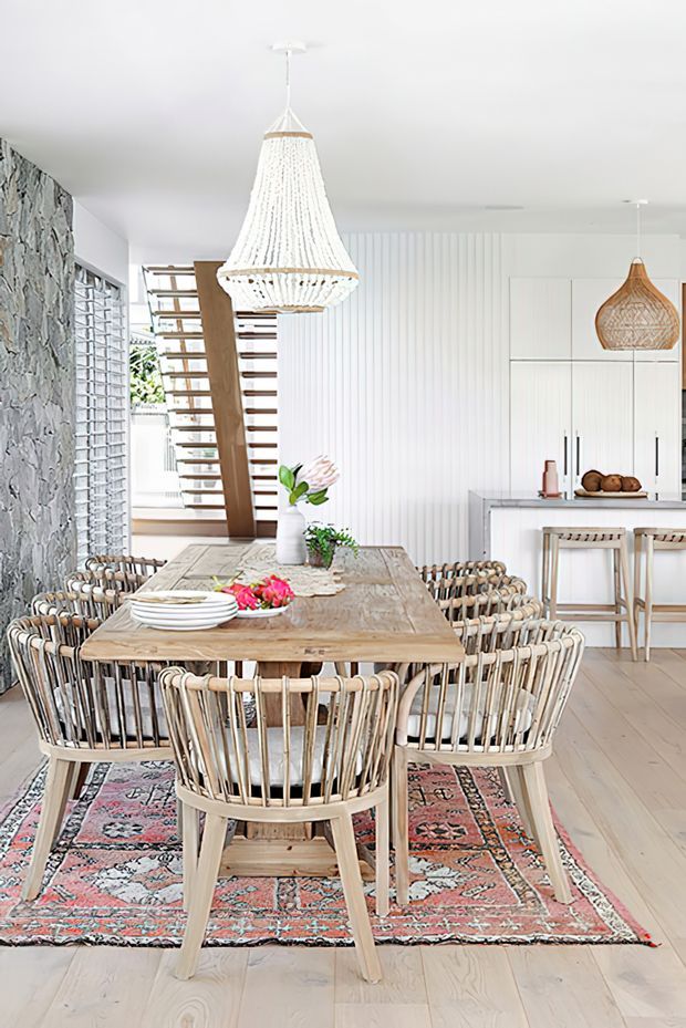 Bright open-plan dining and kitchen interior with a large rustic timber dining table surrounded by woven wooden chairs, a patterned area rug, woven pendant lights, white vertical wall panelling, and a visible open-riser timber staircase. The kitchen includes white cabinetry, a pale island benchtop, and timber bar stools. A textured stone feature wall and glass-block window add visual interest.