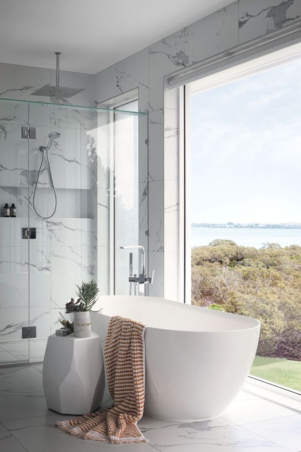 Bright contemporary bathroom featuring a freestanding white bathtub beside expansive glazing, a frameless glass shower with a rain showerhead and handheld fixture, and white marble-look wall and floor tiles with grey veining. The room overlooks a landscaped coastal or waterfront setting, with a small side table, potted plant, and textured towel adding warmth.