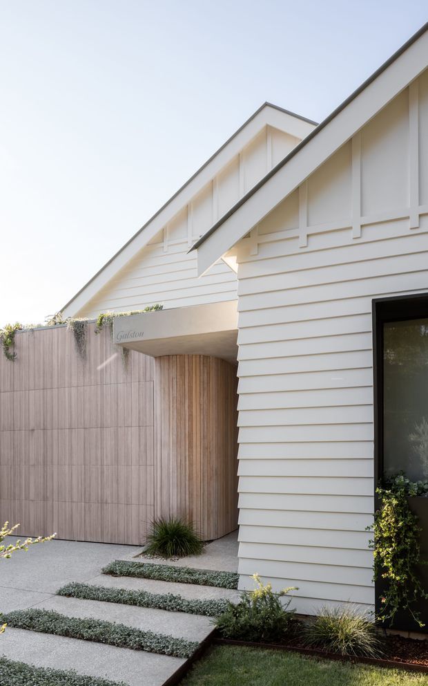 A refined residential facade viewed from the front, featuring white horizontal weatherboard cladding, a steep gabled roof with decorative vertical framing, and a dark-framed entry door. A covered entry recess is lined with vertical timber, opening onto broad concrete stepping slabs bordered by low planting and a tall timber-toned privacy screen. The composition is clean and contemporary with strong gable forms and pale neutral finishes.
