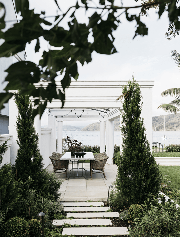 A formal waterfront garden courtyard with a white classical pergola and colonnade framing a dining table and wicker chairs. Large rectangular stepping stones lead through lush layered planting and tall clipped evergreens toward the water, with string lights suspended beneath the pergola. A calm bay, sailboat, hills, and palms are visible beyond the open structure.