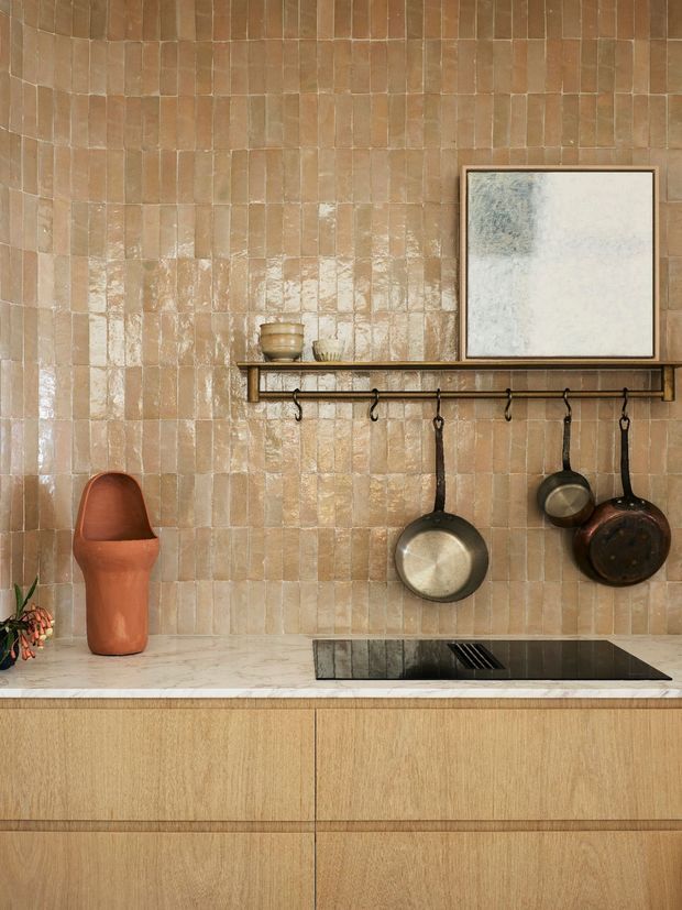 A close-up view of a warm, textural kitchen wall featuring handmade-looking tan ceramic tiles, light timber cabinetry, and a pale stone benchtop. A slim dark shelf with hooks displays hanging cookware, while a black induction cooktop is integrated into the counter. Minimal decorative objects and a framed artwork complete the earthy, refined composition.