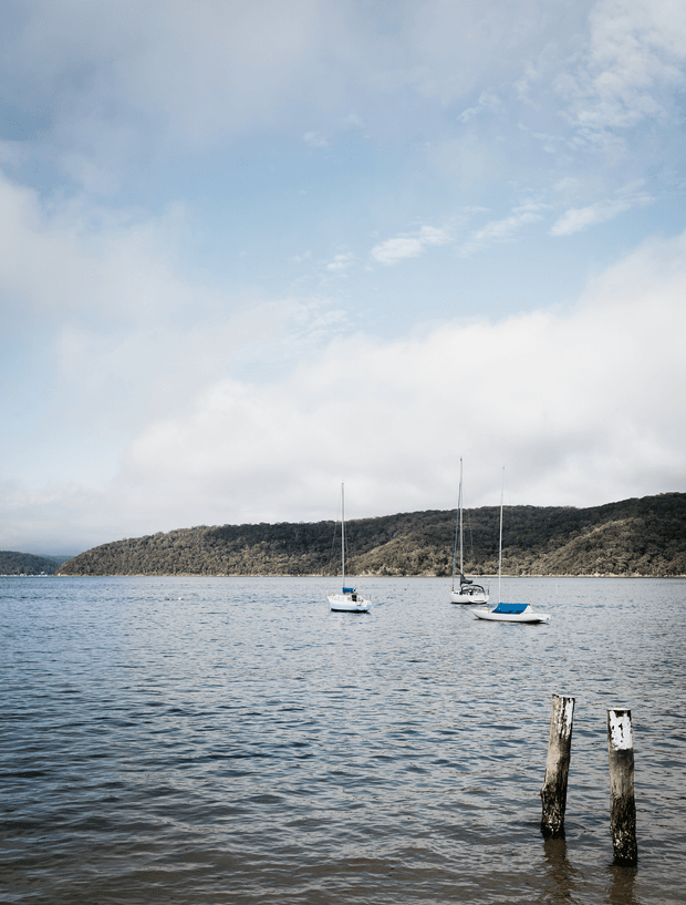 A tranquil waterfront landscape showing calm blue-grey water with three moored sailboats, wooded hills across the bay, and two weathered timber posts in the foreground beneath a partly cloudy sky. No building or architectural space is visible.