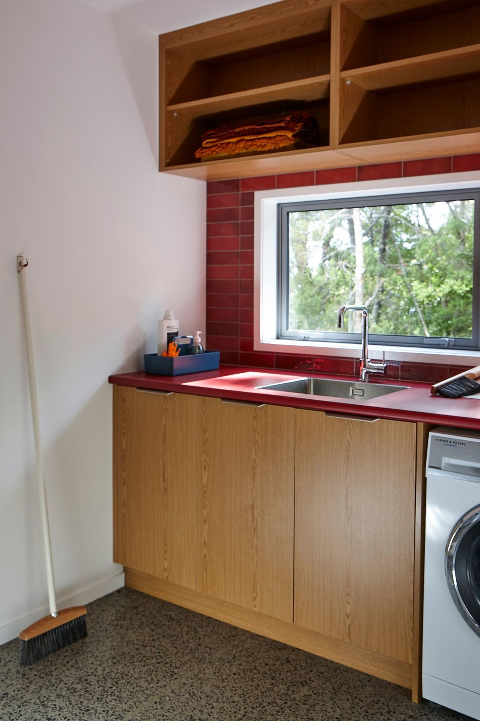The laundry transforms a practical space into a vibrant interior, pairing a red Formica benchtop with oak cabinetry and red tiled splashbacks. A large window frames views into the surrounding New Zealand native forest.