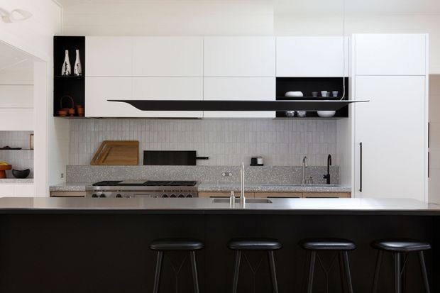 Contemporary kitchen with a long black island and four black stools, white full-height cabinetry, a grey speckled stone benchtop, white textured tile splashback, integrated appliances, and a slim black open shelf feature. The space is clean-lined and monochromatic with strong black-and-white contrast.