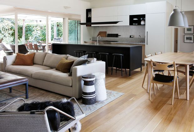 Open-plan contemporary living, dining and kitchen interior with white built-in cabinetry, a dark island, timber flooring, extensive garden-facing glazing, a large sectional sofa and timber dining setting. The bright, relaxed space combines warm natural materials with a clean modern fit-out.