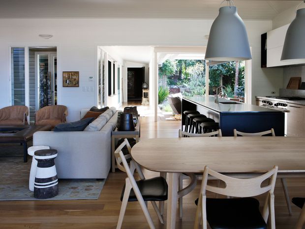 Open-plan coastal-style home interior combining a dining area, living room, and kitchen. A light timber dining table with curved chairs occupies the foreground, while a pale sectional sofa and armchairs define the living area. The kitchen features a dark blue island with integrated seating, pale cabinetry, stainless appliances, and large pendant lights. White wall panelling, timber flooring, abundant glazing, and views to a leafy garden create a bright, relaxed space.
