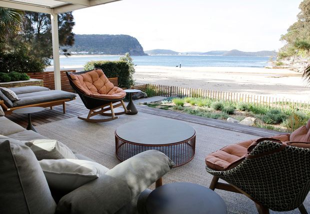 Open-plan covered outdoor lounge overlooking a sandy beach and calm bay. The space features timber decking, a large neutral outdoor rug, low contemporary coffee tables, upholstered lounge seating, leather sling chairs, and a simple overhead pergola structure. Coastal vegetation and a low fence separate the terrace from the beach.