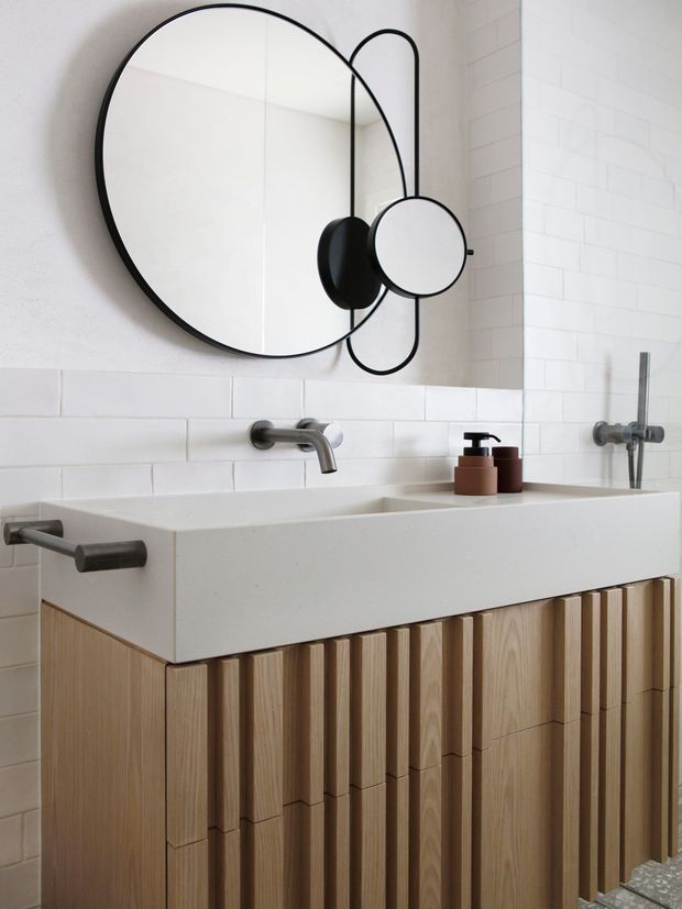A compact contemporary bathroom featuring a wall-mounted white rectangular basin, brushed metal wall-mounted tapware, white rectangular subway tiles, and a vertically ribbed light timber vanity with integrated storage. A large round black-framed mirror with a smaller circular magnifying mirror is mounted above the basin. Neutral finishes and warm timber detailing create a clean, refined look.