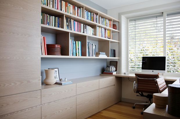 A bright, compact home office with extensive built-in light timber shelving and lower storage cabinetry. The shelving is filled with books and display objects, while a timber desk sits beside a large window with white plantation shutters. A computer and leather office chair complete the workspace. The room has warm timber flooring and a calm, highly organised feel.