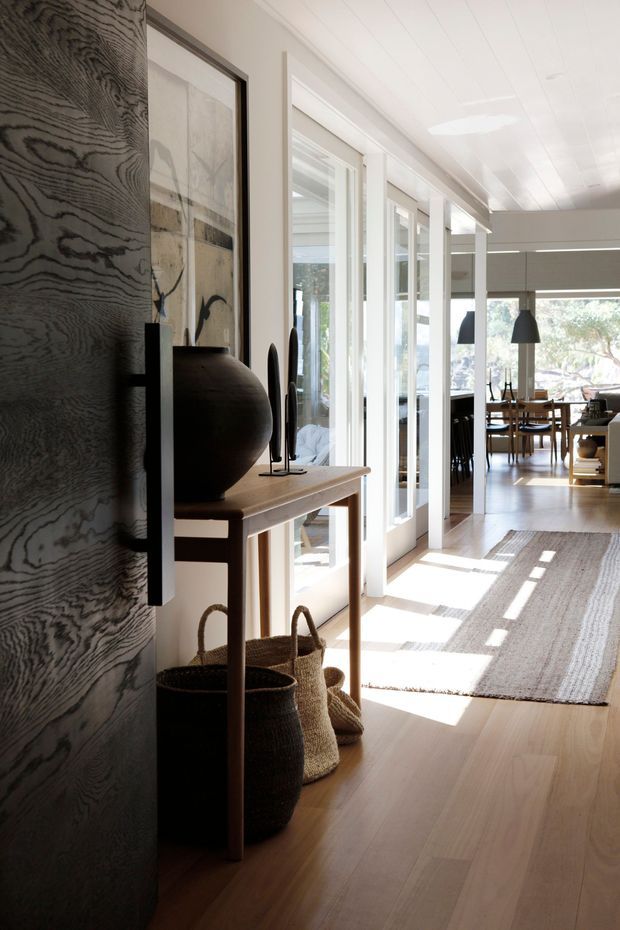 A bright, refined residential hallway or entry passage with warm timber flooring, white-framed glazed doors and partitions, and a slim timber console table styled with dark sculptural decor and woven baskets. The corridor opens toward a dining area with a timber table and pendant lights. A large textured dark wood-grain wall or door forms a strong feature surface in the foreground, while sunlight creates pronounced linear shadows across the floor and rug.