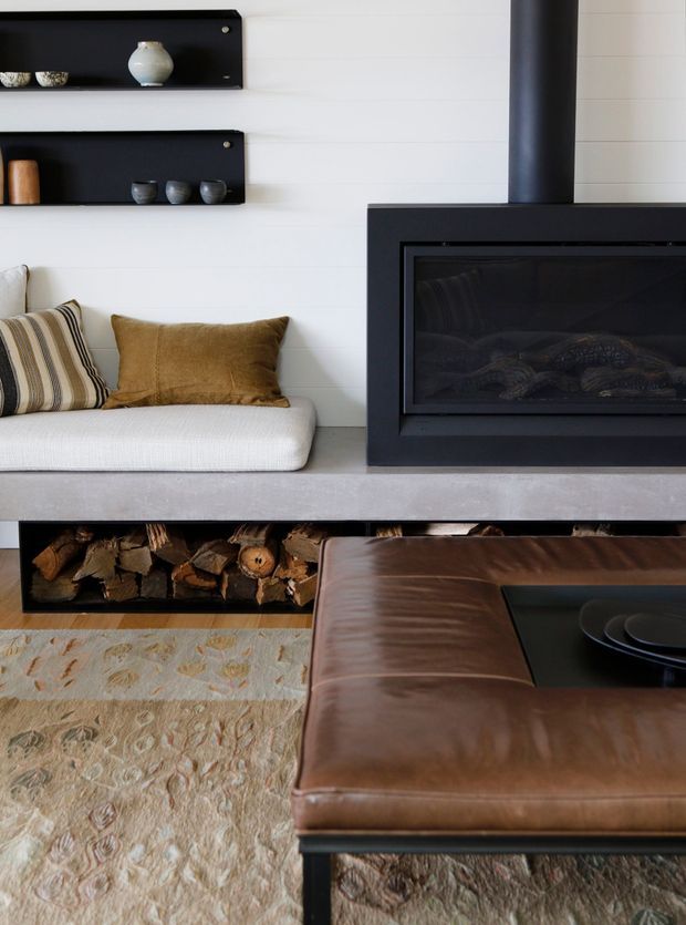 A stylish living room centred on a sleek black fireplace with a matching black flue, set against white horizontal wall panelling. Built-in open shelving displays small ceramics, while a low white upholstered bench with striped and brown cushions sits beside the fireplace. A timber log-storage niche runs beneath the hearth, and a large brown leather ottoman or coffee table occupies the foreground over a textured neutral rug.