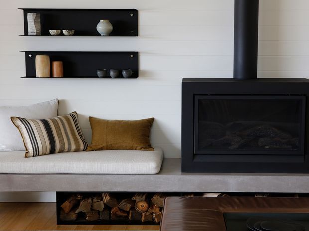 A contemporary living room featuring a built-in black fireplace with a tall flue, a pale upholstered window seat or sofa, timber flooring, and minimalist black wall shelves displaying ceramics. A recessed firewood storage niche sits beneath the concrete-look hearth, with warm neutral cushions and part of a dark leather coffee table visible in the foreground.