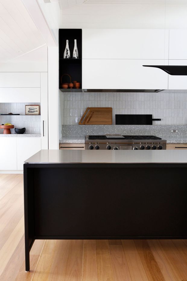 Bright contemporary kitchen featuring a large black island with a pale stone benchtop, white flat-panel cabinetry, a grey terrazzo-like benchtop, vertically stacked white textured tile splashback, black rangehood and appliances, and light timber flooring. Open shelving displays decorative objects, while a partial adjacent dining or living area is visible to the left.