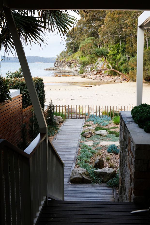 View from a shaded residential entry or terrace looking down a timber boardwalk through a landscaped coastal garden toward a sandy beach and calm bay. The scene includes dark timber decking and railings, a stone retaining wall, native-style planting, and a simple white structural frame, with dense coastal vegetation and rocky shoreline beyond.