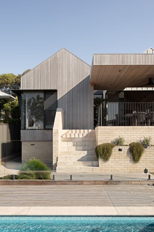 Street-facing exterior photograph of a contemporary residential house beside a swimming pool. The home features pale vertical timber cladding, a prominent gabled volume, dark-framed glazing, a covered upper terrace with timber soffit, pale brick retaining walls, and broad exterior stairs. Low ornamental planting and pool paving occupy the foreground.