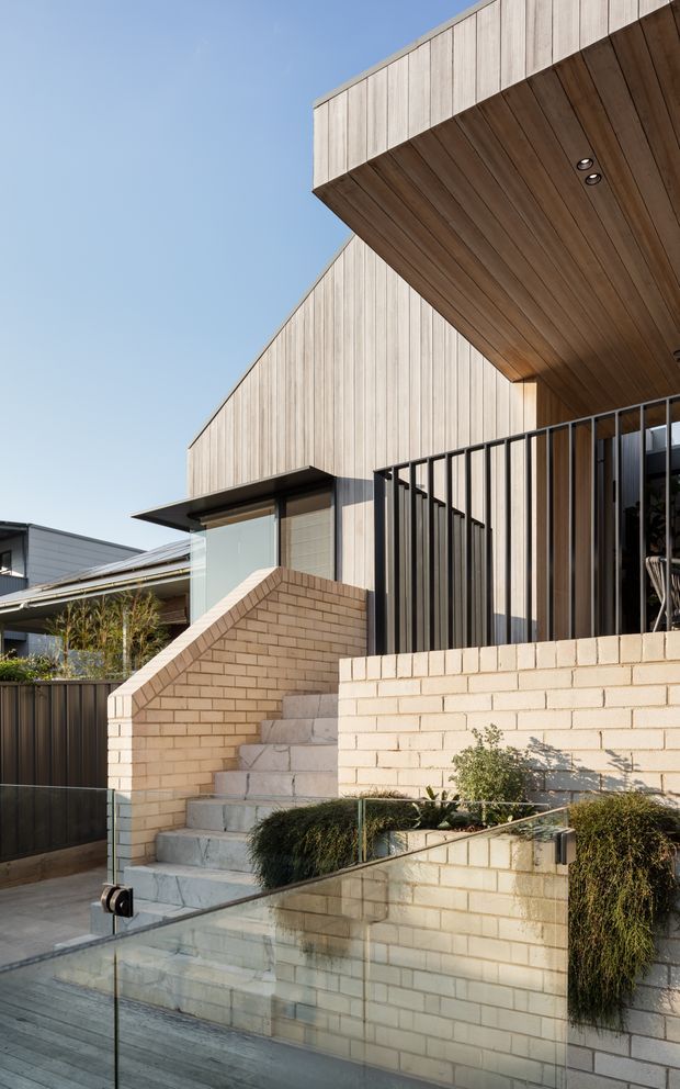 A contemporary multi-level residential exterior viewed from the street, featuring pale vertical timber cladding, a warm timber-lined projecting soffit, cream brick walls, black metal balustrades, broad concrete steps, and clear glass side railings. Landscaped planting borders the stepped entry and a neighbouring modern home is visible in the background.