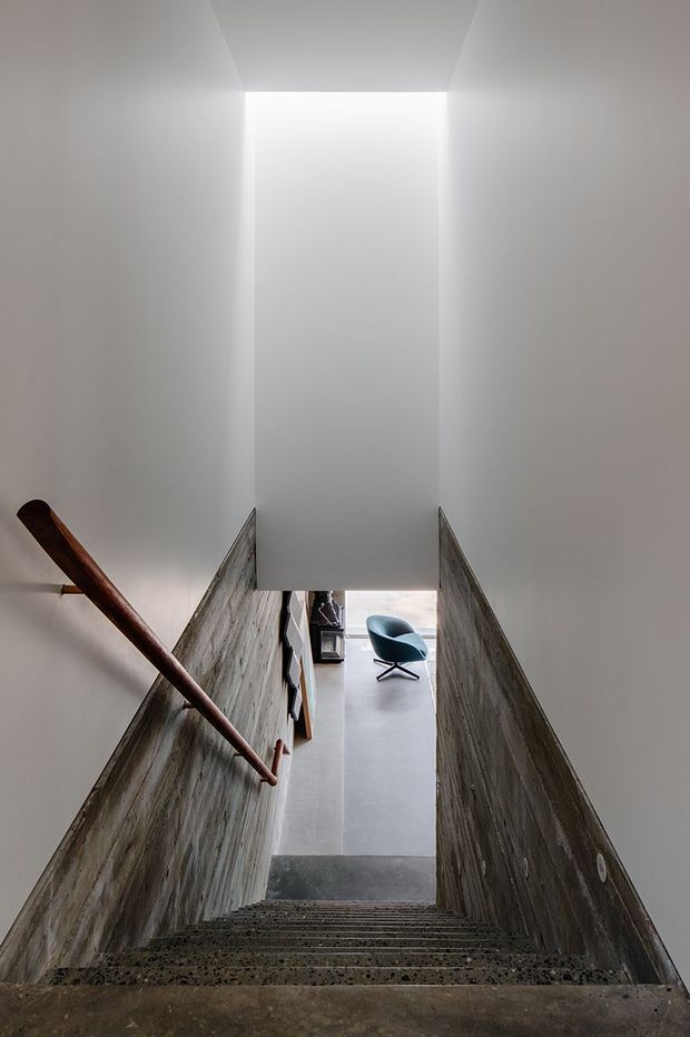 A narrow, minimalist stairway descends toward a small sitting area. The stair walls combine smooth white upper surfaces with dark textured concrete or stone lower walls, and a simple dark timber handrail runs along the left side. Natural light enters from the lower level, where a teal lounge chair is visible on a smooth grey floor.